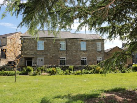 A stone building with white framed windows behind a garden and lawn with trees at St Lawrence Rest in Warkworth