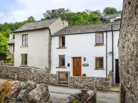 A stone cottage with white walls wooden door and windows surrounded by stone walls and trees at Cinderbarrow Cottage in Levens