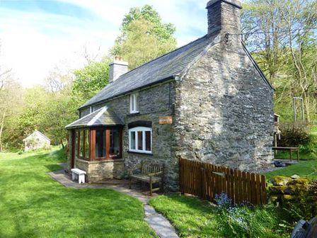 A stone cottage with a wooden bench and fenced garden area surrounded by grass and trees at Glanrafon in Melin-y-Wig near Ruthin