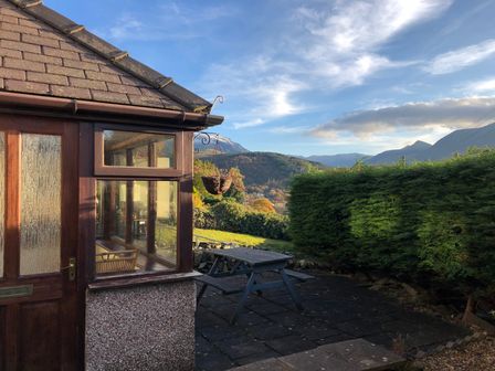 A patio area with a wooden picnic table outside a house with glass windows and a door surrounded by hedges and mountains in Haulfryn Bethesda