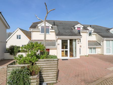 An exterior view of a house with a glass front door and windows on a paved driveway with a wooden planter containing plants at The Beach House in Porth near Newquay