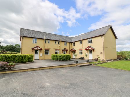 A two story yellow building with multiple doors and windows surrounded by paved area and greenery at Red Kite Cottage in Howey near Llandrindod Wells