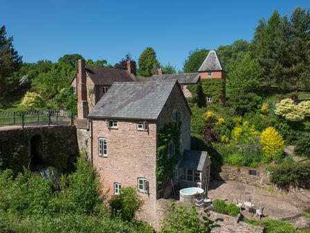 A stone cottage with a slate roof and patio area surrounded by trees and bushes at Mill Cottage in Hanley William near Tenbury Wells