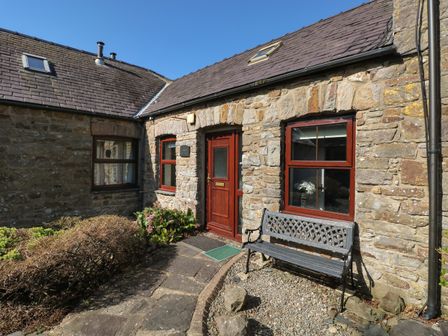 Exterior stone wall of a cottage with a red wooden door and windows and a black metal bench outside at Corner Cottage in Broad Haven