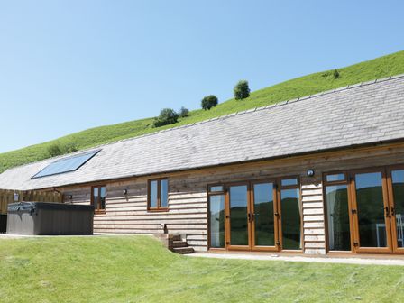 A wooden building with glass doors and windows beside a grassy hill and hot tub at 2 Beacon View Barn in Beguildy near Knighton