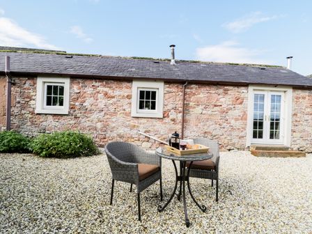 A stone cottage exterior with two windows and French doors a small round glass table with a coffee tray and two wicker chairs on gravel at Faraway Cottage in Kirklinton