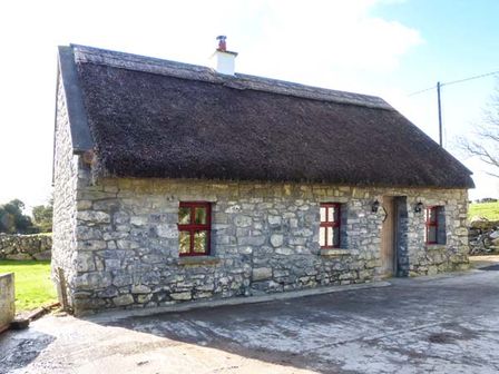 A stone cottage with a thatched roof and red window frames in a rural setting at The Well House in Kinvara County Galway