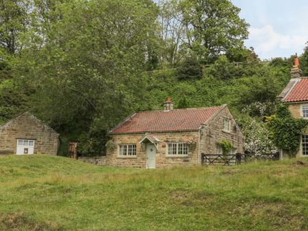 A stone cottage with a tiled roof and hanging flower baskets near a small stone outbuilding and surrounded by grass and trees at Quoits Cottage in Beck Hole near Goathland