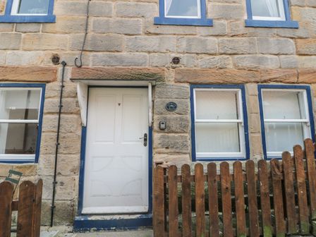 Stone exterior wall with a white door and three windows with blue trim and a wooden fence in front at Grimes Nook Staithes