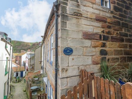 A stone cottage on a narrow cobblestone street with a wooden fence and a sign reading Grimes Cottage in Staithes