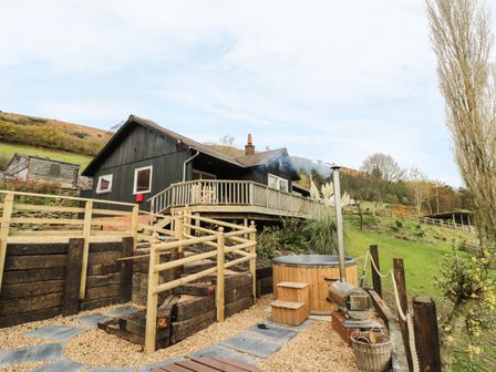 An outdoor area with wooden steps and railing leading to a deck attached to a black house with a hot tub and stove on a gravel surface at Middle Hivron in Bleddfa