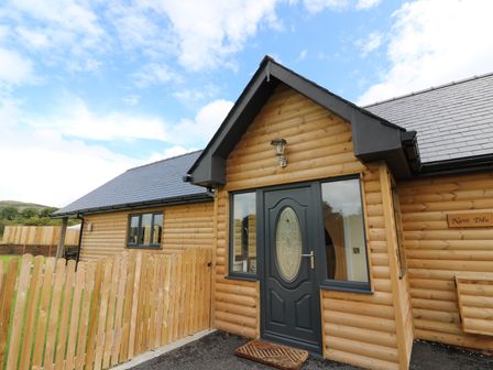 The exterior entrance of a wooden cabin with a black door and windows and a wooden picket fence at Caban Nant Ddu in Rhayader