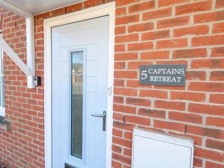 A white front door with a narrow vertical window and a doorbell on a red brick wall with a sign reading 5 Captains Retreat Christchurch