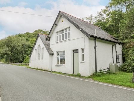 A white house with slate roof next to a road surrounded by trees at Is Y Deri in Harlech