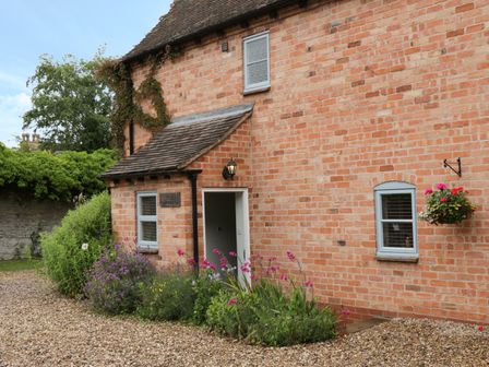 A brick house with gray windows and an open door surrounded by plants and flowers at Pebworth Cottage in Pebworth near Stratford-upon-Avon