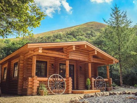 A wooden log cabin with wagon wheels on the porch and trees around at Wilderness Lodge in Stiperstones