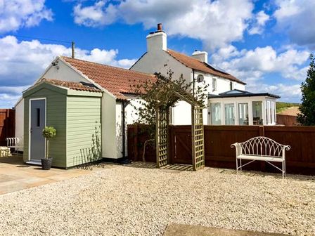 A backyard with a small garden shed a wooden archway a white metal bench and a house with a conservatory at Wold Cottage in Hunmanby