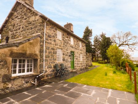 The exterior of a stone house with a green door a paved patio area two chairs and a grassy garden with trees at Ffynnon Gower in Llangower near Bala