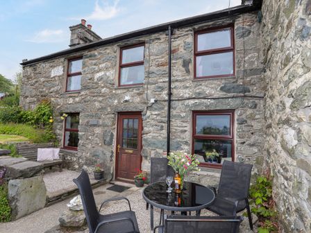 A stone cottage exterior with four windows a wooden door outdoor patio table with chairs and flowers at Hendy Cottage in Llanbedr near Harlech