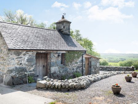An exterior view of a stone cottage with a wooden door and a small stone wall with planters outside at Y Becws in Llanbedr near Harlech
