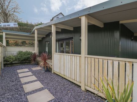 A covered porch area with wooden railing and stepping stones on gravel leading to a dark green corrugated metal building at Y Felin - Moana in Borth near Aberystwyth