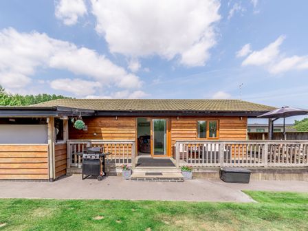 A wooden exterior of a lodge with a patio having a grill two flower pots and an umbrella on a table at Kingfisher Lodge in Brandesburton