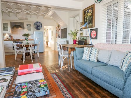 A living room with a blue sofa and wooden coffee table with books next to an open kitchen with a round dining table and white chairs at Bronwen Cottage in Conwy