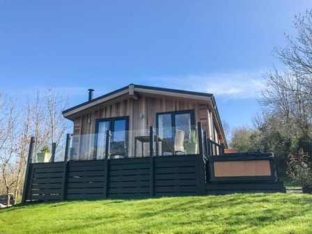A wooden cabin with glass railing on a deck and a hot tub on the side surrounded by grass at Crook Lodge in Allithwaite near Grange-over-Sands