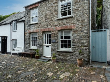 A stone cottage with a white door and windows on a cobblestone street with potted plants near the entrance at Cobble Cottage in Boscastle near Tintagel