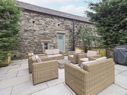 An outdoor patio with wicker sofas and chairs arranged around a small table next to a stone building with a green door and windows at Yew Cottage in Backbarrow