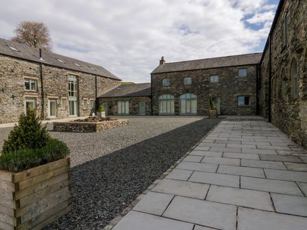 A courtyard with stone buildings surrounding a gravel area and a paved walkway at Rowan Cottage in Backbarrow near Newby Bridge