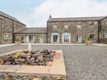 An outdoor courtyard with a stone water fountain in front of a stone building with multiple doors and windows at Lime Cottage in Backbarrow near Newby Bridge