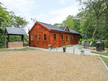 A wooden cabin with multiple windows and doors next to a paved patio with outdoor furniture and a fire pit at Acorn Lodge in Saxmundham