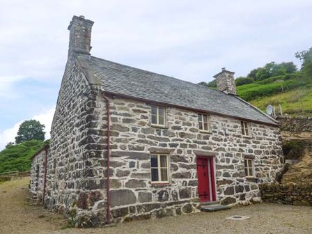 A stone cottage with a red door and multiple windows in a rural setting at Tyn Llwyn in Llanelltyd near Dolgellau