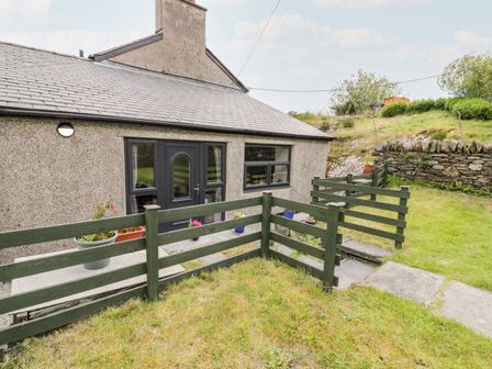 The exterior of a single-story property with a grey door and window beside a small fenced garden with grass and potted plants at Bryn Alun in Tanygrisiau near Blaenau Ffestiniog