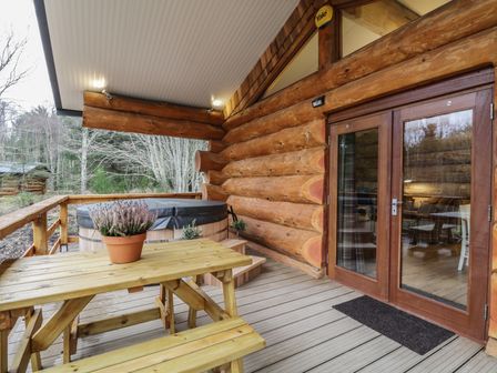 A wooden porch with a picnic table, potted plant, hot tub, and double glass doors at Heron Lodge in North Kessock