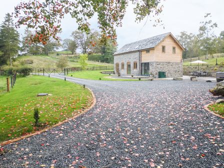 A stone and wood barn house with gravel driveway and green lawn surrounded by trees at Tynddol Barn in Llanbadarn Fynydd