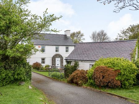 A white house with a stone arch doorway and slate roof near bushes and a metal gate at Cwm Farm in Trearddur Bay