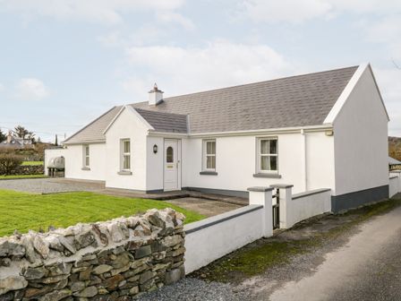 A white single-story house with a grey roof and a stone wall in front at At the Crossroads in Louisburgh County Mayo