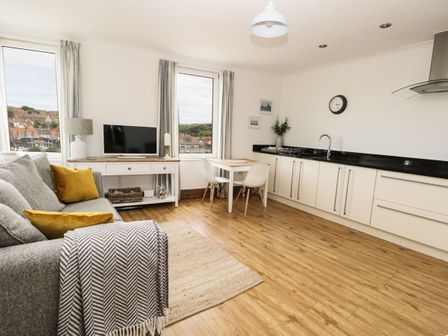 A living area with a gray sofa and yellow cushions a dining table with two white chairs a TV on a wooden console and kitchen cabinets with a black countertop at Marina View in Whitby