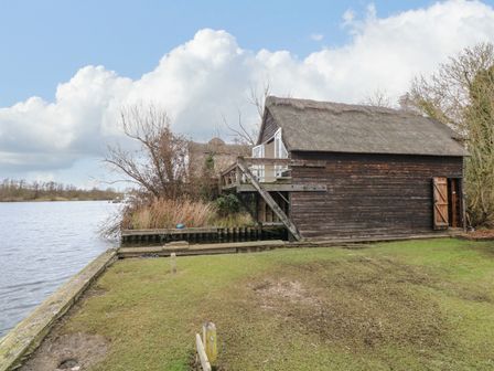 A wooden boathouse with thatched roof by the water with grassy land in front at Cygnus Boathouse in South Walsham