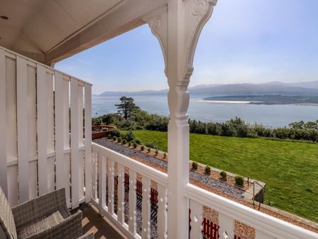 A balcony with white railing and wicker chairs overlooking a garden and water with hills in the distance at Menai View near Menai Bridge