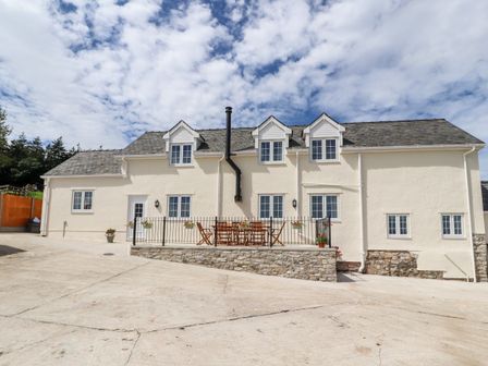 A cream colored house with a stone patio and wooden chairs under a partly cloudy sky at Delfryn Llannefydd near Denbigh