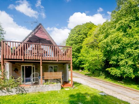 A wooden A-frame house with a balcony and a patio next to a grass lawn and a tree-lined road at Brookside in Honicombe Manor near Gunnislake