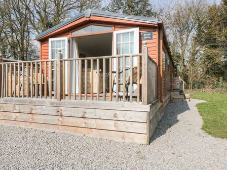 A wooden cabin with an open door and a deck with chairs surrounded by gravel and trees at Broad Down in Berrow