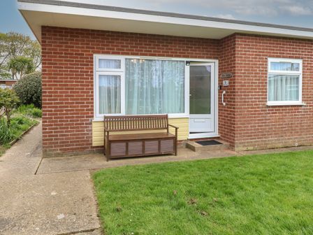 A brick house with a white door and windows a wooden bench on the concrete path and a lawn in front at En-Casa Mundesley