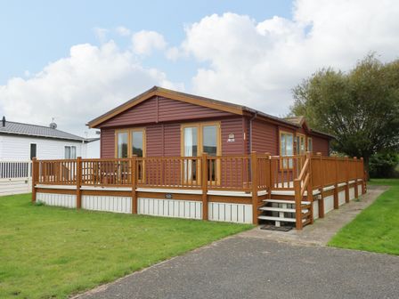 A brown single-story cabin with wooden deck and railing on a grassy plot at Jolie in Hunstanton