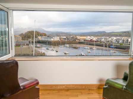 Two chairs facing a large window with a view of a harbor with boats and buildings at The Captain's Penthouse in Porthmadog