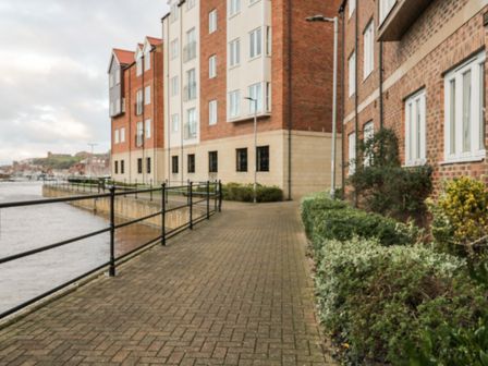 A paved walkway along a waterfront with brick buildings and railing at Anchors Down in Whitby