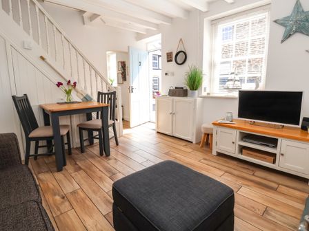 A living room with a wooden table and two chairs a grey ottoman a television on a cabinet and an open door at Sandy Shell Cottage in Conwy